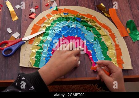le mani del bambino incollano la carta colorata su un arcobaleno di cartone. compiti della scuola da fare a casa. Orizzontale Foto Stock