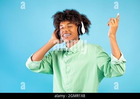 Gioiosa teen nera che ascolta musica in cuffie, chiudi gli occhi e rilassati sullo sfondo blu dello studio Foto Stock