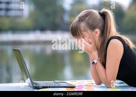 Giovane donna sesaurita che lavora dietro un computer portatile sdraiato in un parco estivo all'aperto. Problemi con il concetto di lavoro remoto. Foto Stock