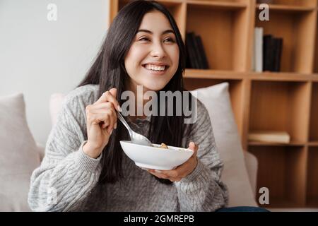 La giovane donna emozionata mangia cereali mentre guardava la tv su un divano a casa Foto Stock