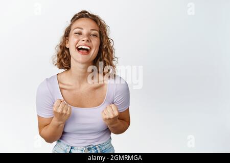 Ragazza bionda allegra e motivata ridendo, sorridendo e guardando felice, pugni clenching incoraggiati, in piedi su sfondo bianco Foto Stock