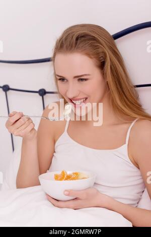 Allegra donna caucasica bionda che mangia frutta fresca sul letto Foto Stock
