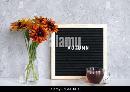 Testo autunnale su tavola nera, bouquet di fiori arancioni in vaso e tazza di tè sul tavolo contro parete di pietra grigia. Modello per cartolina, messaggio di saluto Foto Stock