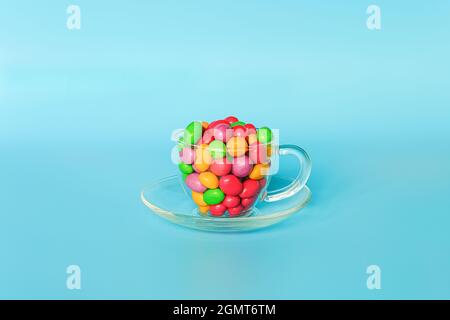 Fagioli caramelle colorati glassati. Tazza di vetro sul piatto piena di colorati cioccolatini a forma di bottoni su sfondo blu con spazio per la copia. Tè dolce di concetto pa Foto Stock