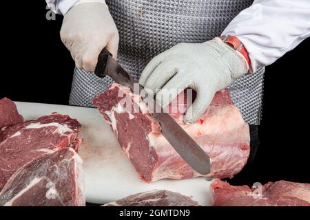 Primo piano di pezzi di carne macinata e le mani maschili del macellaio in guanti speciali che tagliano con il coltello. Carne di maiale o di manzo sul tavolo del macellaio. Lavoratore in whit Foto Stock