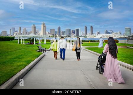 Vista posteriore di una donna con lunghi capelli scuri e di altre persone che si dirigono verso l'area del Coronado Ferry Landing con lo splendido skyline di San Diego, California, in lontananza Foto Stock