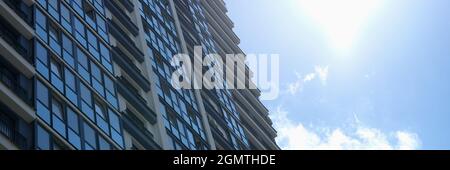 Edificio a più piani con molte finestre di vetro che si erigono sullo sfondo di cielo blu e sole Foto Stock