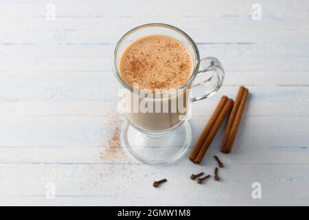 Tè piccante con latte in una tazza di vetro con bastoncini di cannella e chiodi di garofano su sfondo blu grigio chiaro Foto Stock