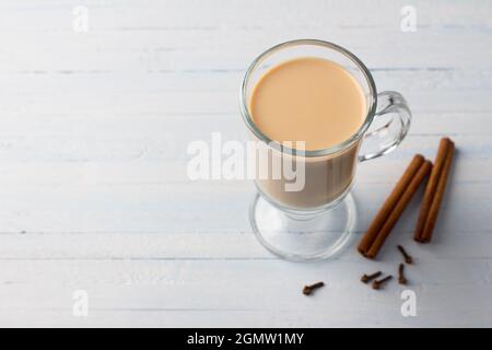 Tè piccante con latte in una tazza di vetro con bastoncini di cannella e chiodi di garofano su sfondo blu grigio chiaro Foto Stock