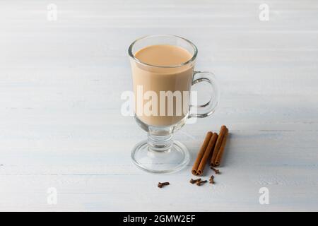 Tè piccante con latte in una tazza di vetro con bastoncini di cannella e chiodi di garofano su sfondo blu grigio chiaro Foto Stock