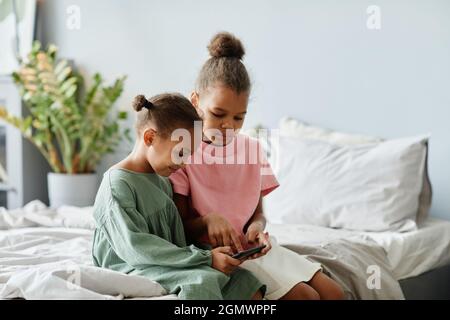 Ritratto di due simpatiche ragazze afroamericane usando lo smartphone insieme mentre si siede a letto in una stanza accogliente, spazio copia Foto Stock