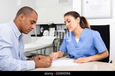 Giovane gentile latino cosmetologo medico lavorando con il cliente maschile in clinica, compilando la forma medica prima della procedura .. Foto Stock