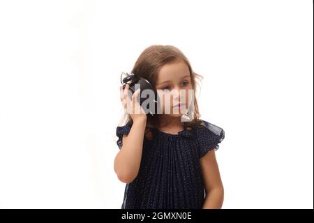 Una bella ragazza curiosa di 4 anni in un abito blu scuro tiene una sveglia vicino al suo orecchio e ascolta attentamente il suono delle frecce, Foto Stock