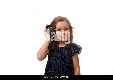 Adorabile bambina di 4 anni in un abito blu scuro tiene una sveglia vicino all'orecchio e ascolta attentamente il suono, sorride guardando la macchina fotografica Foto Stock