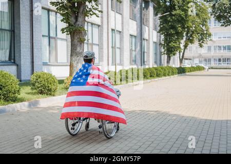 vista posteriore del soldato disabili, avvolto nella bandiera degli stati uniti, seduto in sedia a rotelle sulla strada urbana Foto Stock