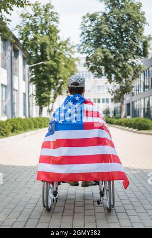 vista posteriore del veterano in sedia a rotelle, avvolta nella bandiera degli stati uniti sul vicolo della città Foto Stock