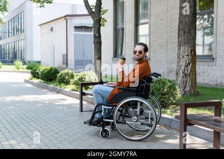 giovane uomo handicappato seduto in sedia a rotelle con computer portatile e drink da asporto all'aperto Foto Stock