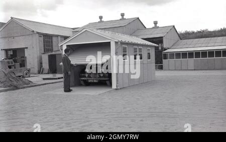 Anni '50, storico, all'esterno su un'ampia area pianeggiante in una zona industriale leggera, un uomo in un vestito che solleva la porta di roll-up di garage prefabbricato, costruito lì per scopi dimostrativi, Witney, Oxford, Inghilterra, Regno Unito. Questi garage o ripari per motori sono stati realizzati con pannelli in calcestruzzo prefabbricato imbullonati insieme in sezioni, con un tetto in metallo ondulato e una porta arrotolante in metallo. Con l'aumento della proprietà delle auto alla fine degli anni '50 e '60, questi garage auto-costruiti o kit in cemento divennero popolari. Foto Stock