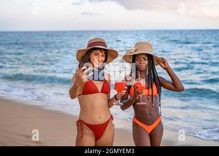 Affascinante giovane donna multirazziale amici in bikini e cappelli prendere selfie su smartphone mentre in piedi con bicchieri di bevande sulla spiaggia sabbiosa contro il mare in estate sera Foto Stock