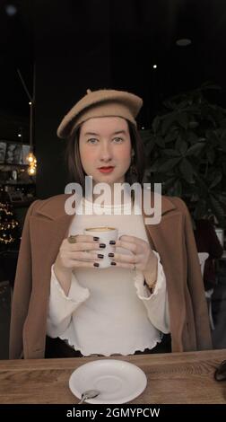 Giovane donna in un bar che tiene in mano una tazza calda di caffè pronto a bere in posa per la macchina fotografica. Autunno Vibes Parigino Girl per 2021. Foto Stock