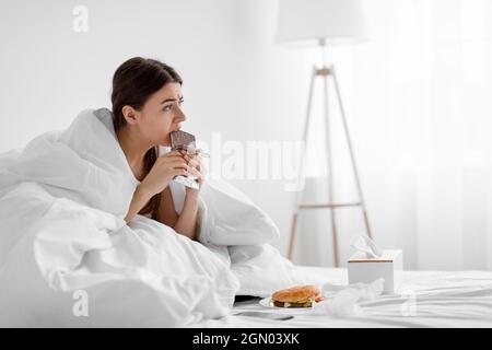 Delusa donna millenaria che mangia la tavoletta di cioccolato a letto con fast food a casa, soffrendo di mangiare troppo Foto Stock