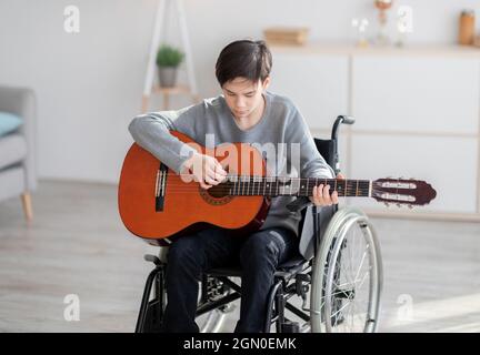 Teen boy disabili focalizzato in sedia a rotelle suonare la chitarra, creando musica a casa. Rimani hobby a casa durante il blocco covid Foto Stock