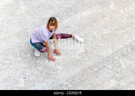 Da sopra tutto il corpo di giovane donna vestita in elegante abbigliamento sportivo e sneaker che fanno esercizio stretching per le gambe su quadrato urbano pavimentato durante la fi all'aperto Foto Stock