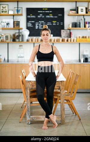Donna sorridente e tranquilla in abbigliamento sportivo in piedi nudi vicino al tavolo e rilassante prima di allenarsi in palestra Foto Stock