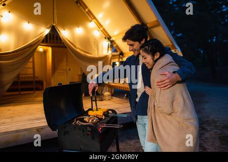 Vista laterale di sorridente cucina interrazziale sulla terrazza glamping Foto Stock