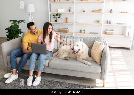 Allegro giovane coppia multirazziale seduta sul divano con computer portatile, stroking loro cane a casa, spazio copia Foto Stock