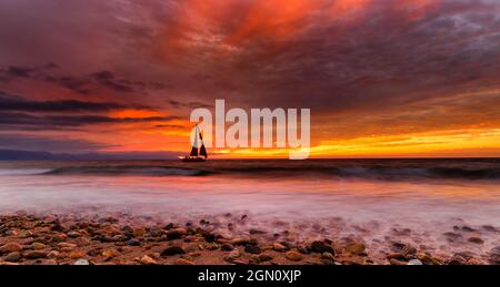 Una barca a vela sta navigando lungo l'oceano con un gregge di uccelli che volano sopra UN cielo colorato di tramonto Foto Stock