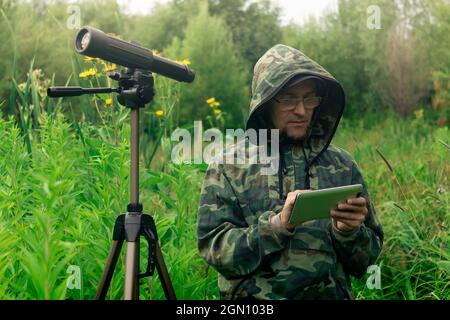 l'ornitologo uomo guarda o scrive le informazioni sulla tavoletta mentre si trova in piedi tra l'erba alta nella zona umida Foto Stock