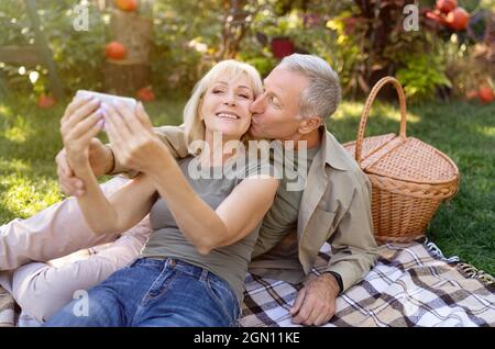 Felice coppia senior prendere selfie su smartphone, fare un picnic e riposare in giardino all'aperto, godendo la natura autunno Foto Stock