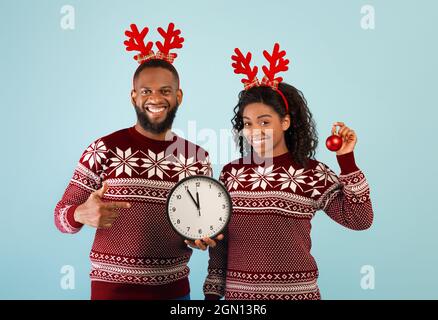 Festeggiamo l'anno nuovo. Sposi allegri neri con orologio che mostra cinque minuti fino a mezzanotte, indossando corna di cervi e maglioni, posando su sfondo blu, Foto Stock