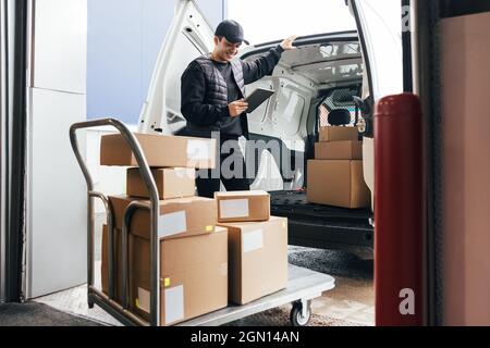 Corriere in uniforme che tiene un tablet digitale che controlla la quantità di pacchi su un carrello mentre si trova in piedi in auto Foto Stock