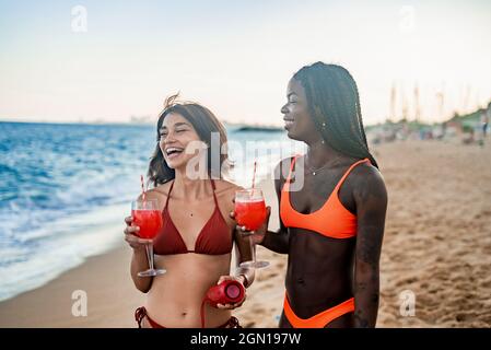 Giovani e diverse amiche in bikini ridono felicemente mentre bevono cocktail rossi e ascoltano musica con altoparlanti audio portatili sulla spiaggia sabbiosa durante le vacanze estive insieme Foto Stock