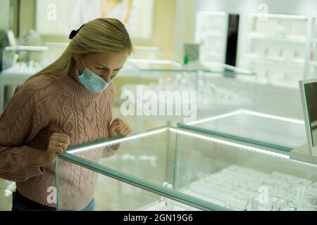 donna in una maschera da coronavirus e con capelli biondi sceglie gioielli nel negozio Foto Stock