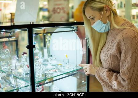 donna in una maschera da coronavirus e con capelli biondi sceglie gioielli nel negozio Foto Stock