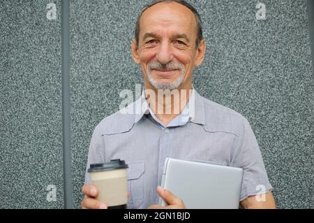 Ritratto maschile del collega più anziano con computer portatile argento e tazza di caffè in attesa di una riunione vicino al muro grigio scuro al mattino, lavoro remoto, caffè Foto Stock