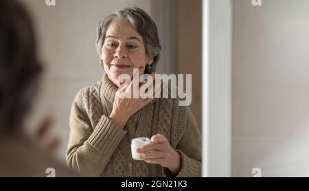 Una donna anziana applica un idratante anti-invecchiamento. Foto Stock