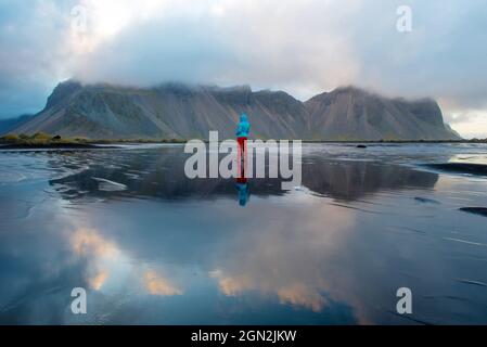 Paesaggio islandese, riflesso di una donna che cammina e del monte Vestrahorn nelle acque dell'oceano Atlantico. Penisola di Stokksnes, Hofn, Icel Foto Stock