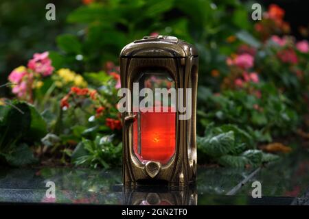 lampada in metallo con candela a fuoco su una tomba con fiori colorati su sfondo sfocato Foto Stock