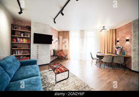 Vista dell'interno di un ampio soggiorno con grande divano blu. Decorazione luminosa di appartamento con libreria, TV, ampia finestra e curpet su pavimento in legno. Concetto di design del soggiorno. Foto Stock