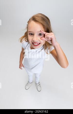 Vista ad alto angolo della bella bambina con capelli lunghi in un abito moderno ed elegante che si posa isolato su sfondo bianco studio. Concetto di infanzia felice. Foto Stock