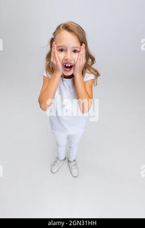 Vista ad alto angolo della bella bambina con capelli lunghi in un abito moderno ed elegante che si posa isolato su sfondo bianco studio. Concetto di infanzia felice. Foto Stock