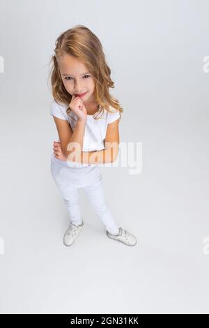 Vista ad alto angolo della bella bambina con capelli lunghi in un abito moderno ed elegante che si posa isolato su sfondo bianco studio. Concetto di infanzia felice. Foto Stock