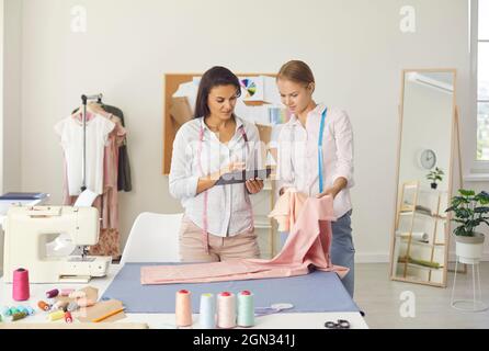 Due donne che possiedono un piccolo lavoro di dressmaking utilizzando il tablet mentre lavorano nel loro atelier Foto Stock