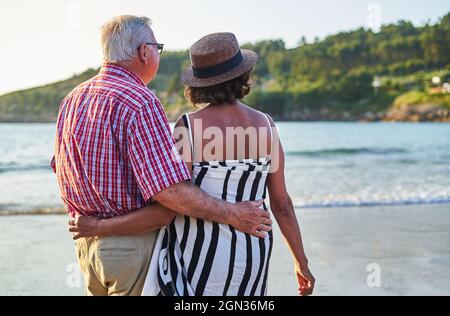 Vista posteriore di una coppia di anziani irriconoscibili in occhiali da sole in piedi su spiaggia di sabbia bagnata e godere di una giornata di sole Foto Stock