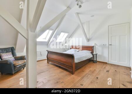 Biancheria da letto su letto in legno contro poltrona retrò e finestre sopra il parquet a casa durante il giorno Foto Stock