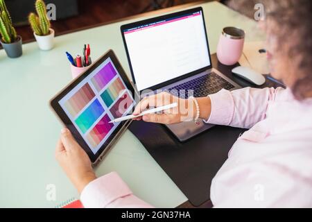Ritagliare lo schermo a sfioramento della donna anziana sul tablet puntando la tavolozza dei colori e parlando durante la chat video sul netbook in ufficio Foto Stock